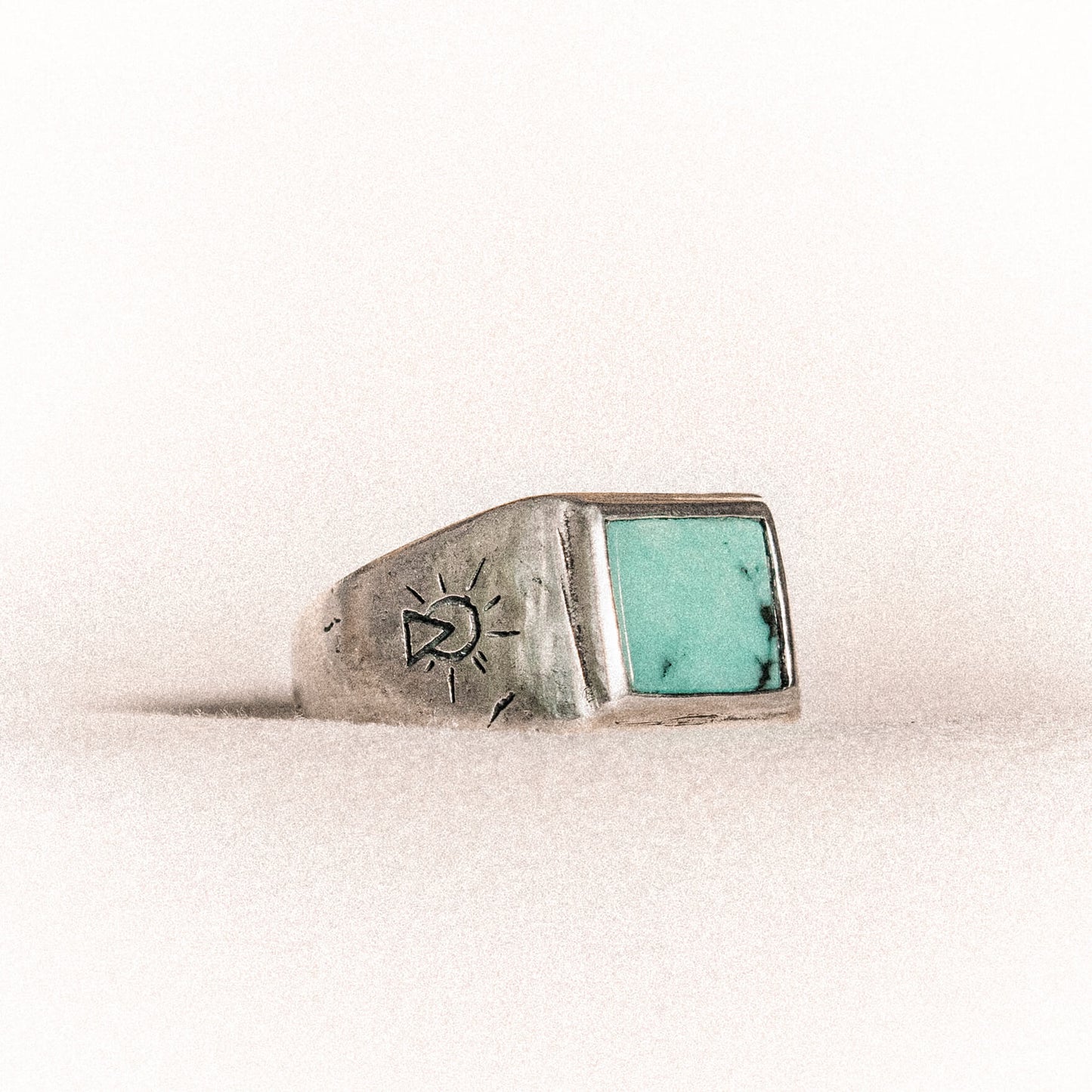 A silver ring with a rectangular turquoise stone and a small sun-like symbol engraved on the side, set against a neutral background.